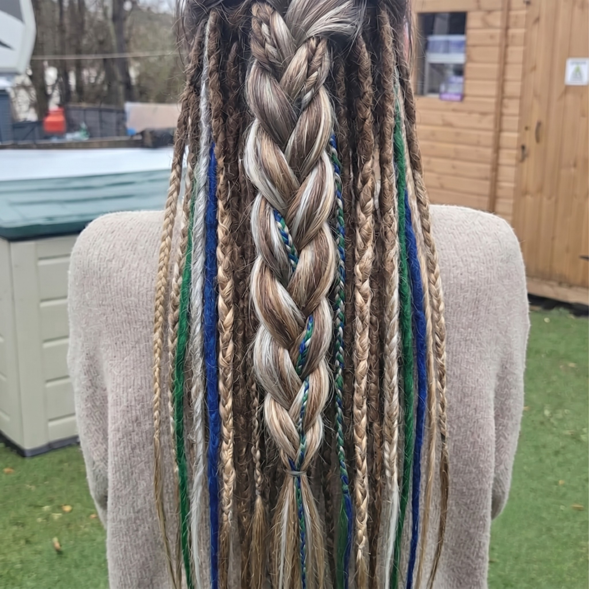 Model wearing brown blonde dreadlock extensions with blue, green, white and silver accents, styled in a braided back view with loose end synthetic dreads.